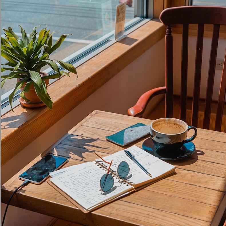 An empty coffee cup and an open notebook sit on a wooden table in a sunny cafe.